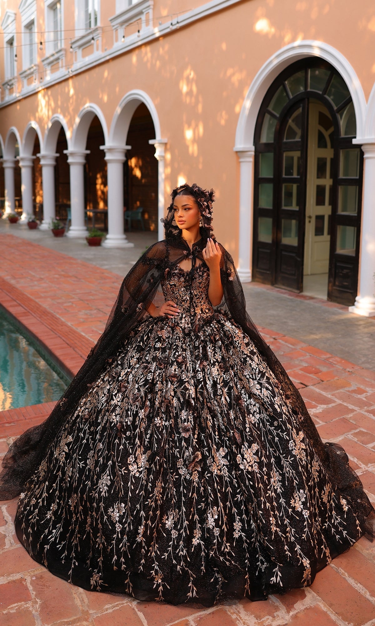 A woman stands by a pool in front of an arched building, wearing the Quinceanera Dress 54320 By Amarra?an ornate off-the-shoulder ball gown with silver embroidery, 3D floral details, and a matching headpiece.